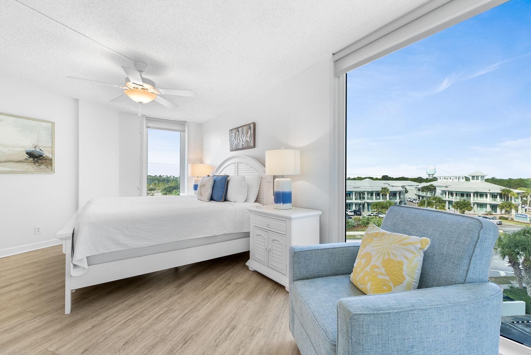 A bedroom with a bed, a chair, a nightstand, a ceiling fan, a window, and a view of buildings.