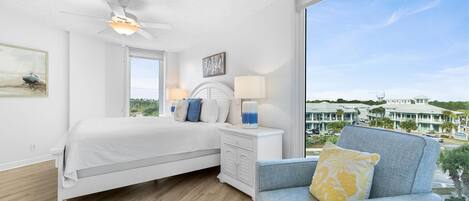 A bedroom with a bed, a chair, a nightstand, a ceiling fan, a window, and a view of buildings.