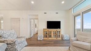 A spacious living room with a large window, a flat-screen TV mounted on the wall, and a wooden cabinet. - Vista Del Mar 402 (Pensacola)