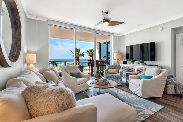 A spacious living room with a large TV, comfortable seating, and a balcony with a view of the ocean.