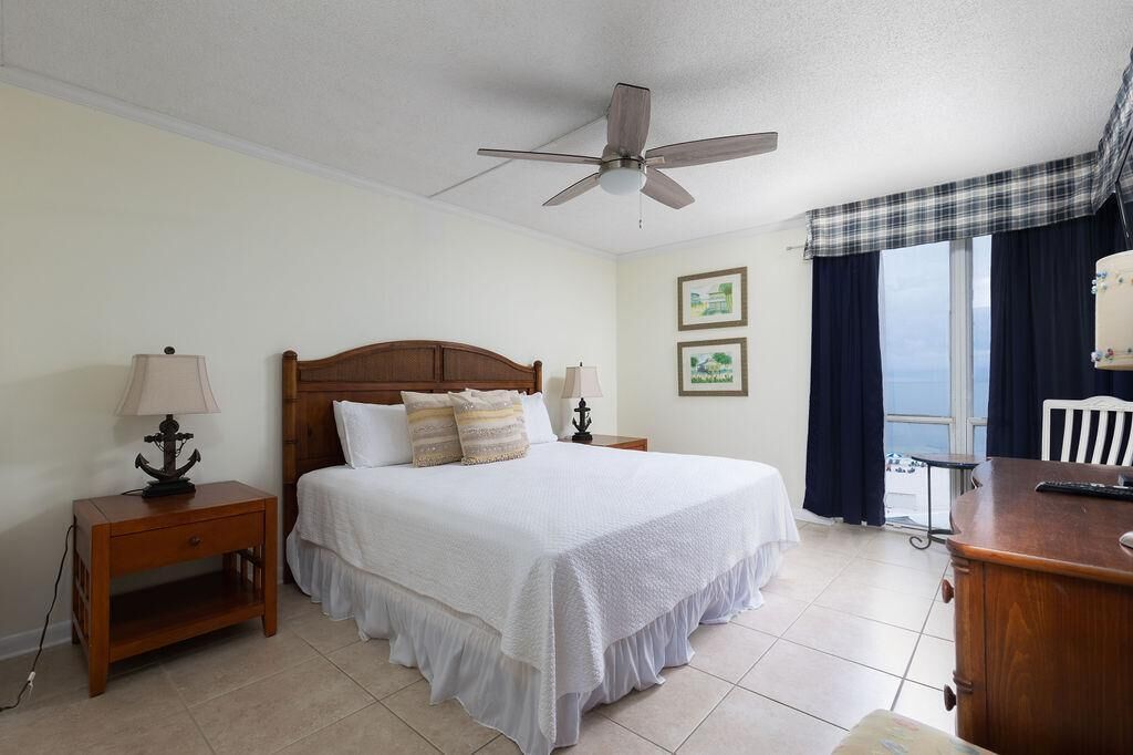 A bedroom with a bed, a nightstand, a desk, a ceiling fan, and a window with curtains.