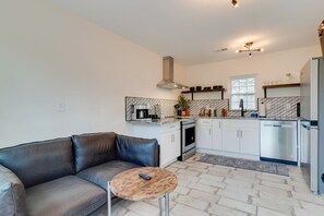 A modern kitchen with white cabinets, a stainless steel refrigerator, and a built-in oven. There is a leather sofa and a small round table with a remote control on it.