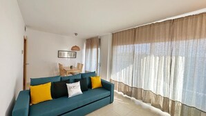 A living room with a teal sofa, yellow and black cushions, a dining table with chairs, and large windows with sheer curtains. - Seaside Getaway: Spacious Apartment with Terrace, Pool & Parking in Salou (Salou)