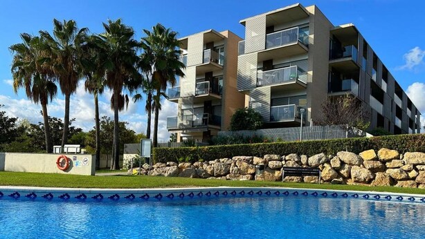 A modern multi-story building with balconies, a swimming pool, palm trees, and a stone wall. - Seaside Getaway: Spacious Apartment with Terrace, Pool & Parking in Salou (Salou)