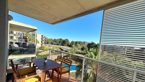A balcony with a table and chairs, overlooking a swimming pool and a landscaped area with trees and a building in the background. - Seaside Getaway: Spacious Apartment with Terrace, Pool & Parking in Salou (Salou)