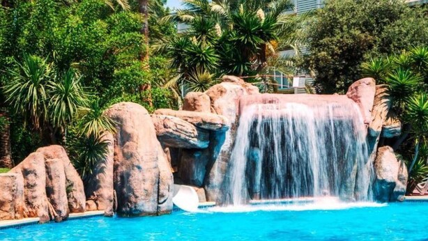 Ein Pool mit Wasserfall, umgeben von großen Steinen und tropischer Vegetation.