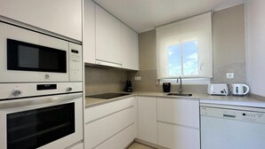 Eine moderne Küche mit weißen Schränken, integriertem Geschirrspüler, Backofen und Mikrowelle, einem Fenster mit Blick und einer Spüle mit Wasserkocher und Toaster.