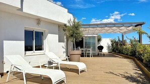 Eine Dachterrasse mit weißen Liegestühlen, einem Tisch und einer Grillgelegenheit.