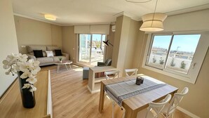 Ein modernes Wohnzimmer mit Sofa, Couchtisch und Esstisch. Große Fenster bieten Blick auf die Stadt.