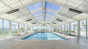 A large indoor swimming pool with a high ceiling and skylights, surrounded by large windows and potted plants. - Perdido Sun 702 (Pensacola)