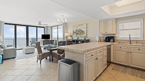 A modern kitchen with a large island, dining area with chairs, and a view of the ocean through large windows. - Lei Lani 607t (Orange Beach)