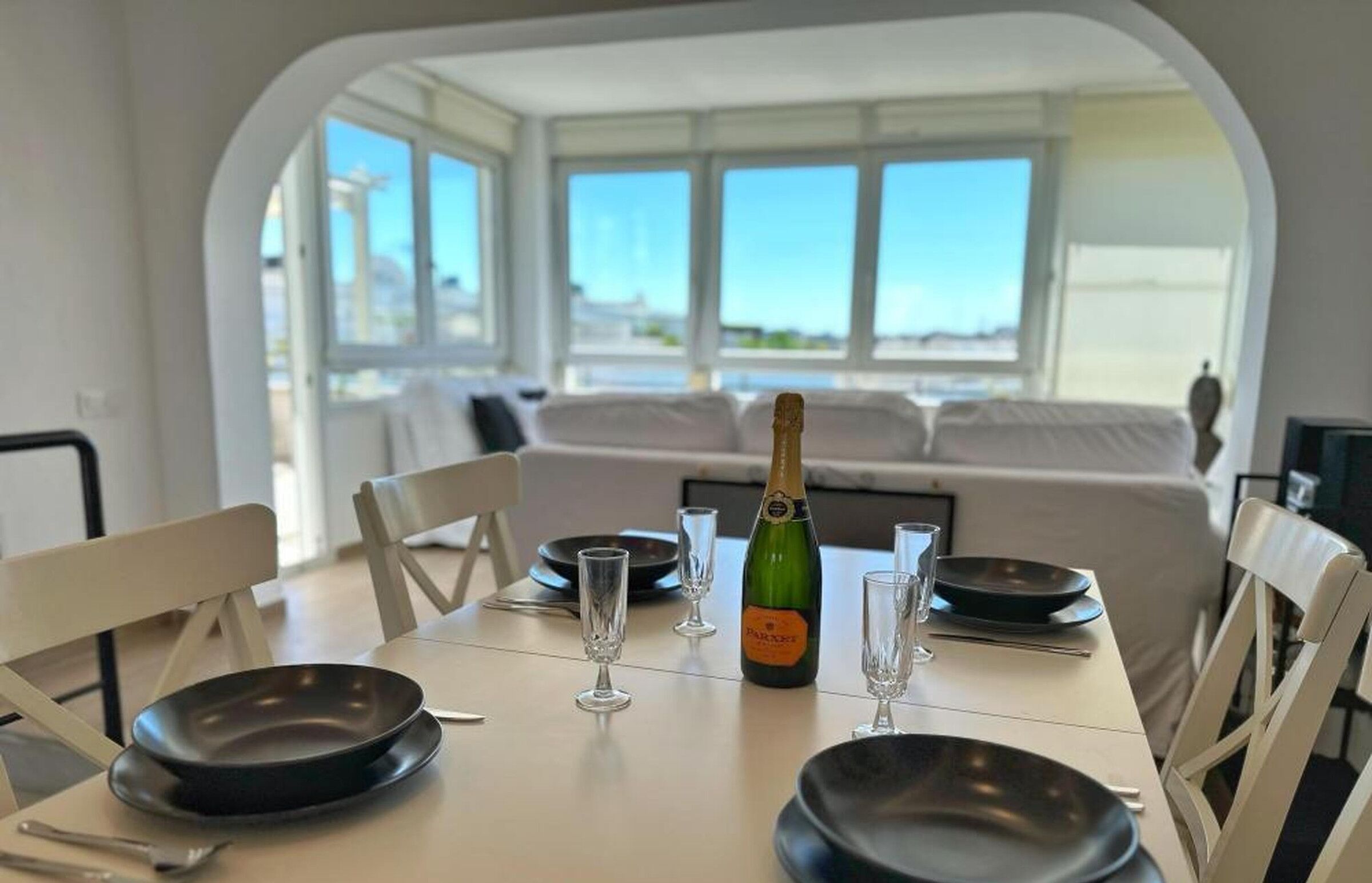 Une table à manger dressée avec des assiettes, des verres et une bouteille de champagne, dans une pièce lumineuse offrant une vue sur la mer.