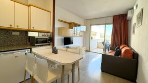 A compact kitchen with a microwave, stove, and cabinets, a dining area with chairs, and a sofa in a living area with a view to the outside. - Cozy Apartment with Pool, 300m from Levante Beach, Free WiFi, Near Port Aventura (Salou)