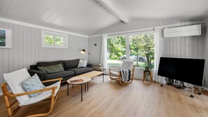 A cozy living room with a sofa, a chair, a coffee table, a TV, and large windows with a view of greenery outside.