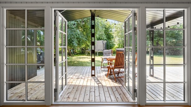 A covered patio with a wooden deck, a table with chairs, and a view of a garden with trees and a grill.