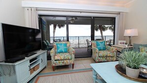A living room with a TV, two patterned armchairs, a sofa, a coffee table, and a view of the beach through a sliding glass door. - Shipwatch A201 (Pensacola)