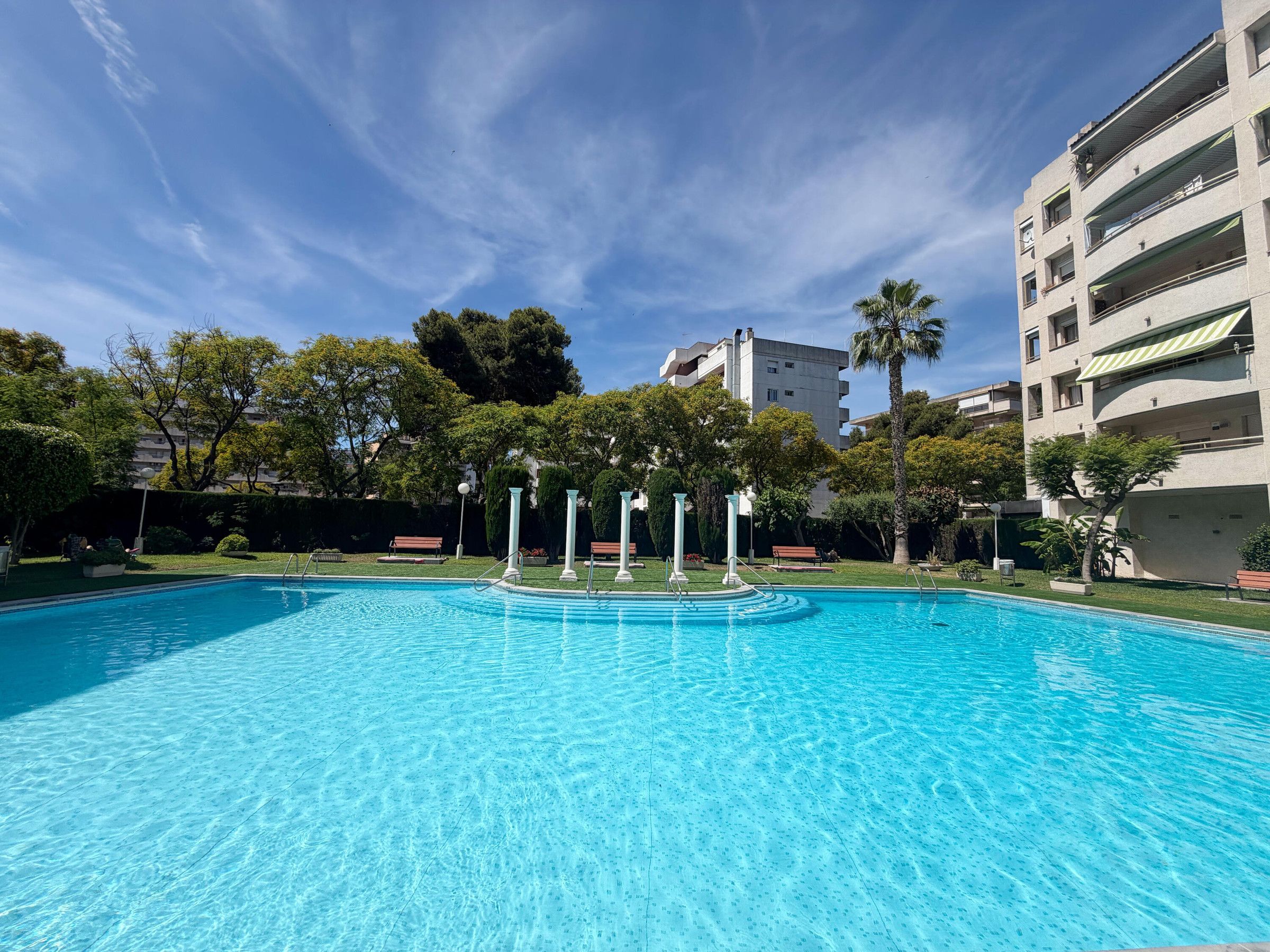 A swimming pool with clear blue water, surrounded by greenery and buildings.