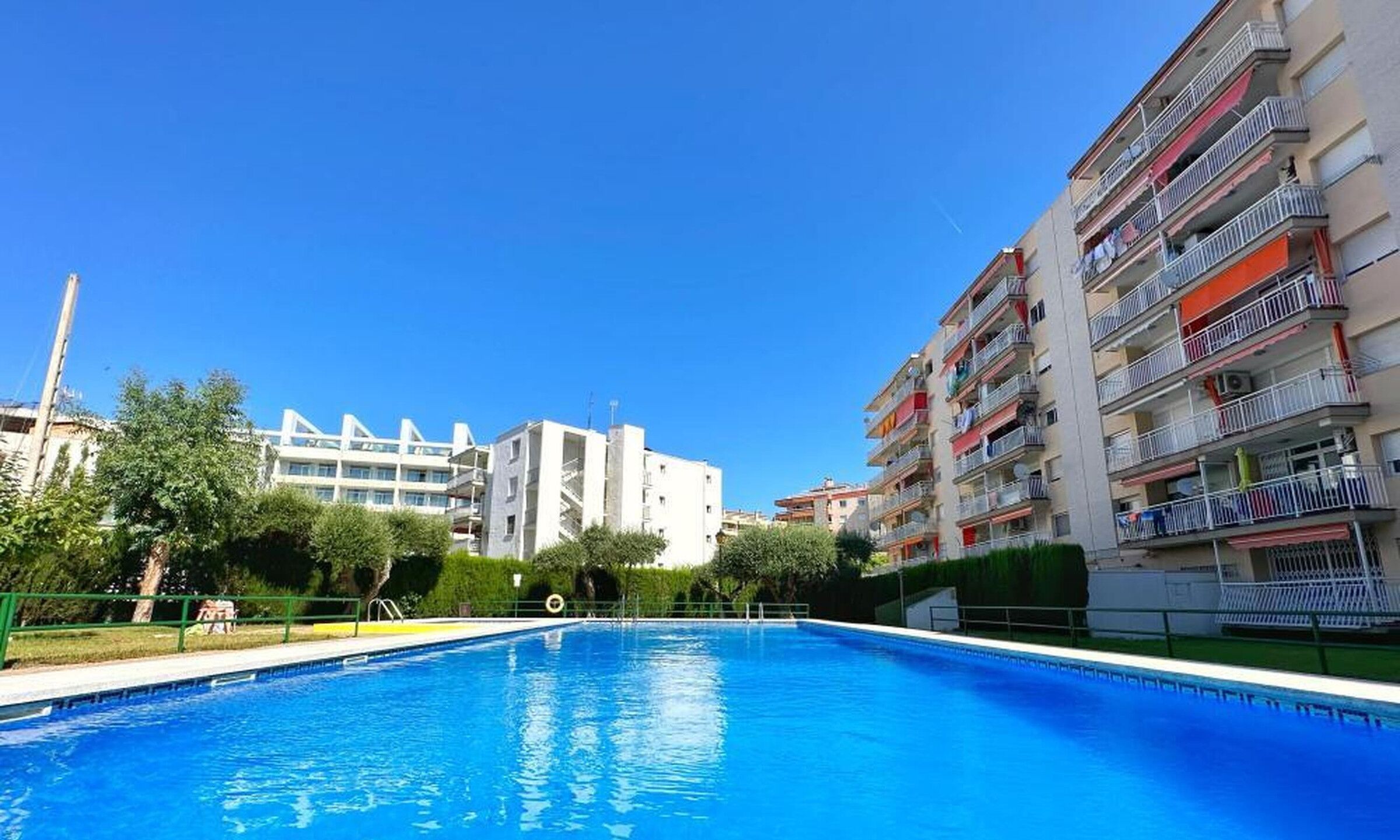Una piscina bajo un cielo azul despejado, rodeada de edificios residenciales con balcones.