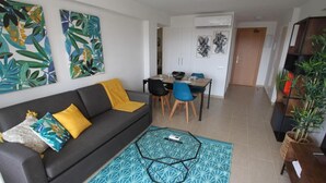 A living room with a sofa, coffee table, and a dining area with chairs and a table. There are two wall-mounted artworks and a door in the background. - Cozy Apartment with Garden, Pool & Parking - Steps from Playa del Llevant! (Salou)