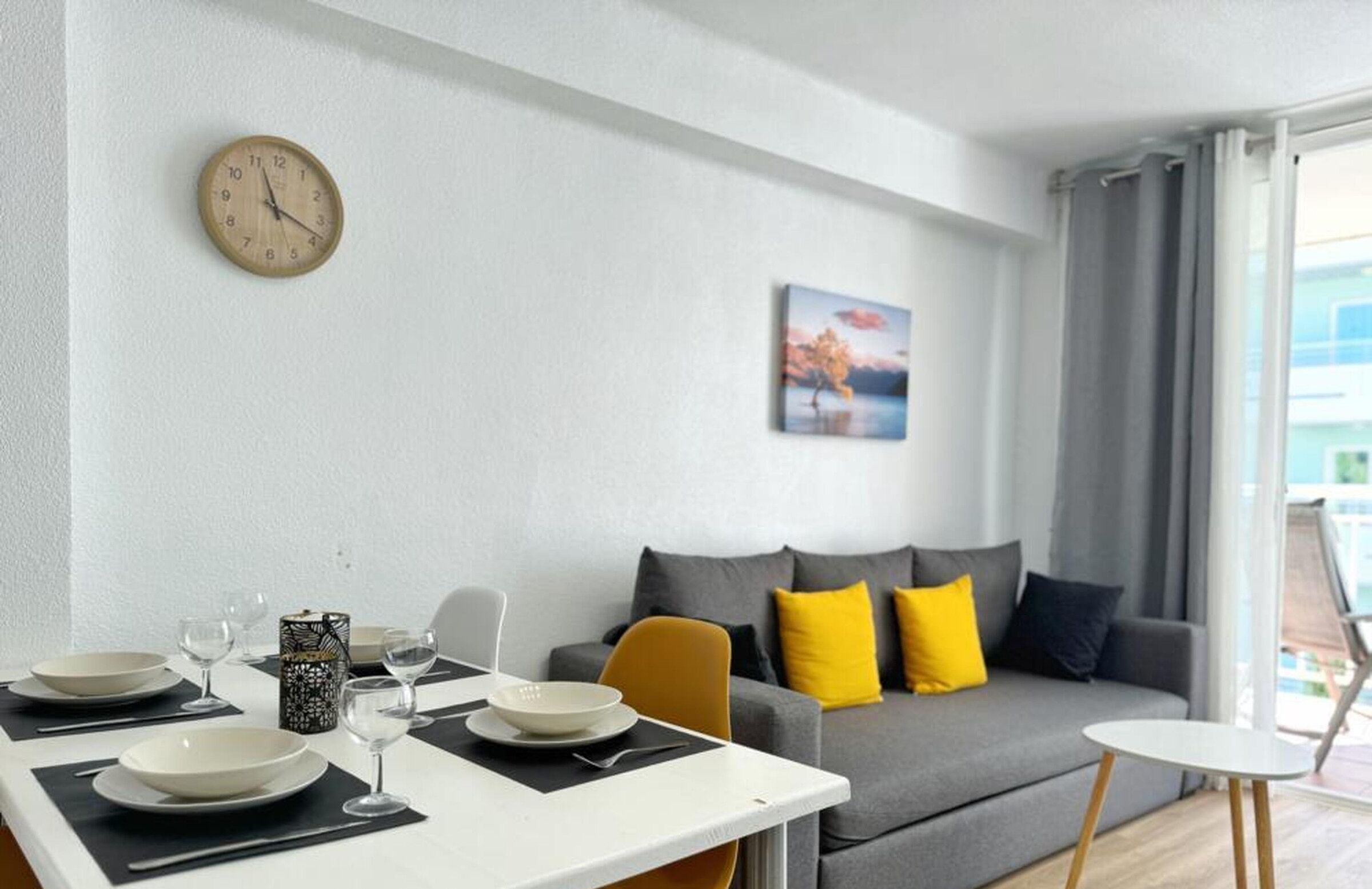 A modern living room with a dining area, a sofa with yellow cushions, a clock on the wall, and a painting.