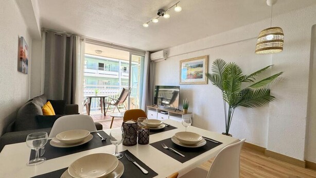 Un comedor con una mesa dispuesta para cuatro personas, un sofá, un televisor y una planta en maceta.