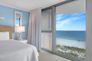 A hotel room with a large window overlooking a beach, a bed with a white comforter, a bedside table with a lamp, and a decorative mirror.