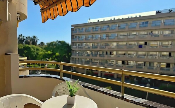 Un balcon avec une table blanche, un pot de fleurs et une vue sur un immeuble à plusieurs étages.