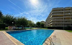 Une piscine sous un ciel bleu dégagé, entourée d’une zone pavée et d’un immeuble à plusieurs étages.