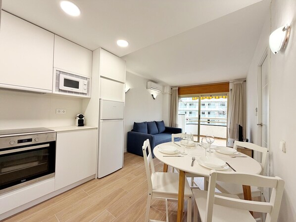Una cocina compacta con electrodomésticos modernos, un comedor con mesa y sillas blancas, y un sofá azul al fondo.