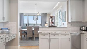 A kitchen with white cabinets, a dining area with wicker chairs, a view of the ocean, and a balcony with a table and chairs. - Indigo West 1104 (Pensacola)