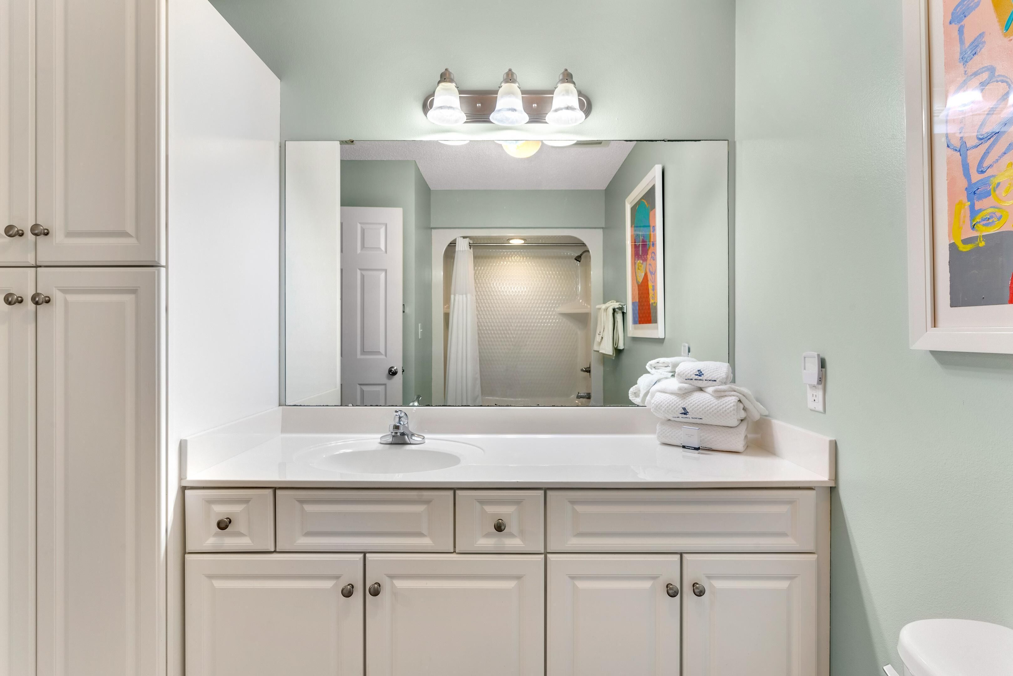 A bathroom with a white vanity, a sink, and a mirror above it. There are towels stacked on the vanity, and a shower area is visible in the background.