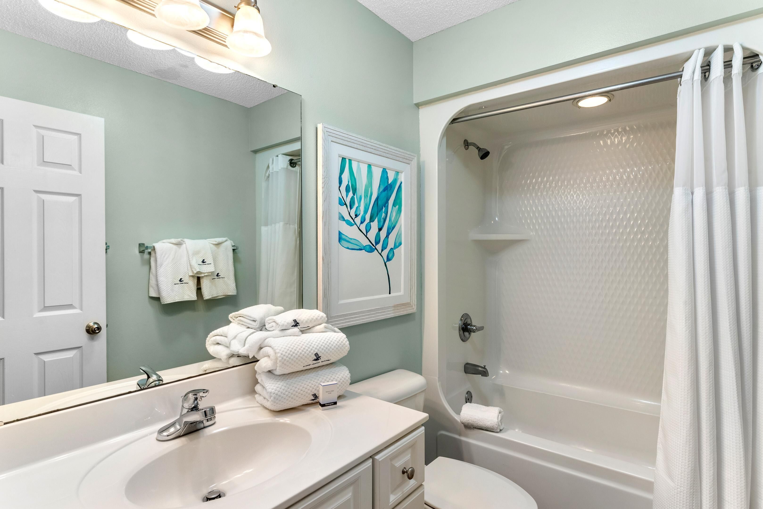 Une salle de bain avec un lavabo blanc, des toilettes, une douche avec un rideau blanc et un miroir orné d’un motif représentant des feuilles.