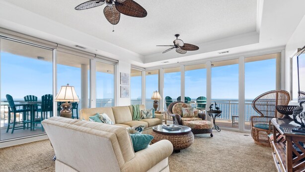 A spacious living room with a large sofa, a coffee table, and a dining area with chairs. There are two ceiling fans and large windows offering a view of the ocean. - La Playa 801 (Pensacola)