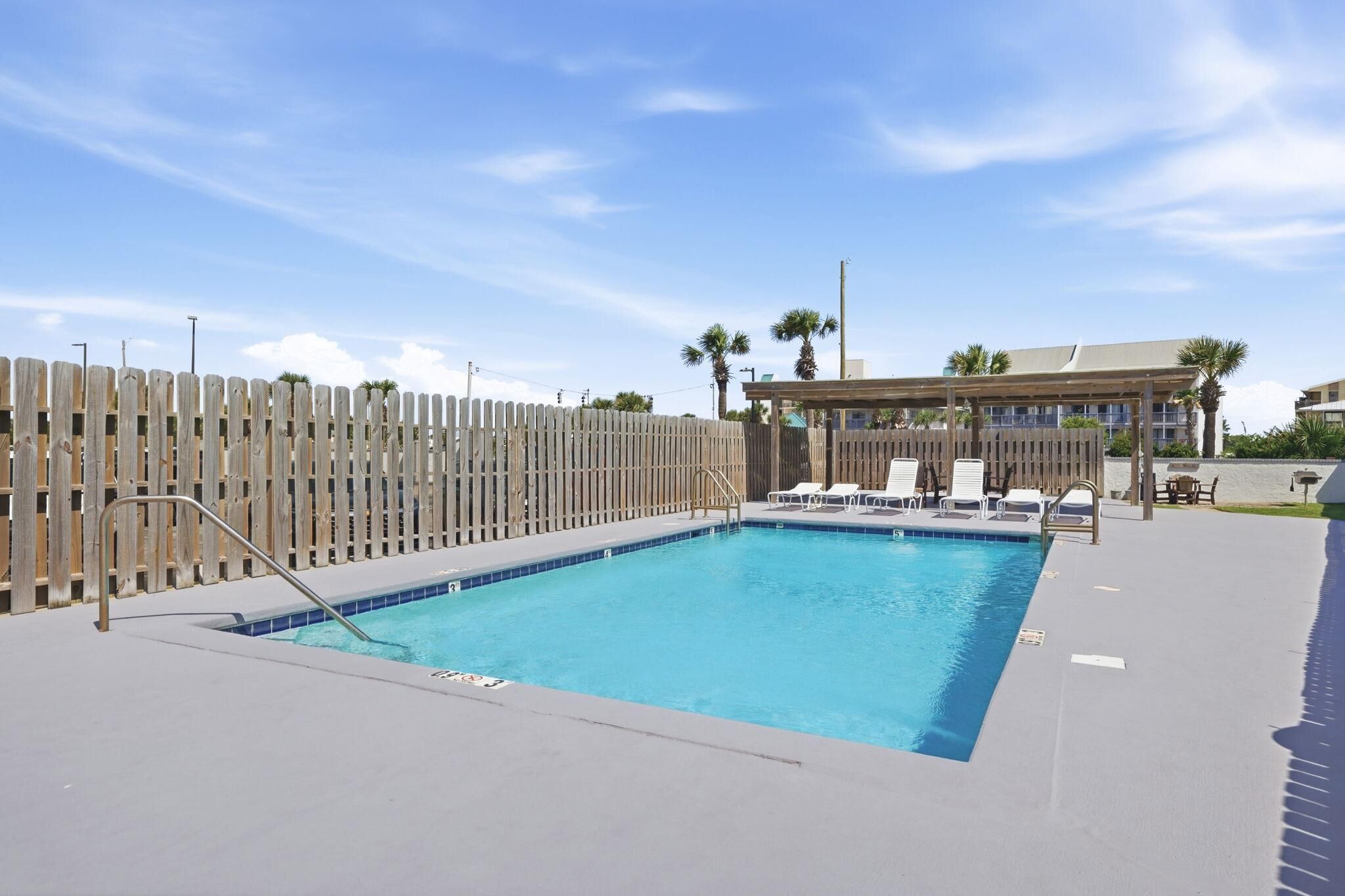 Una piscina rectangular con cerramiento de madera, sillas de lounge blancas y una zona de estar cubierta.