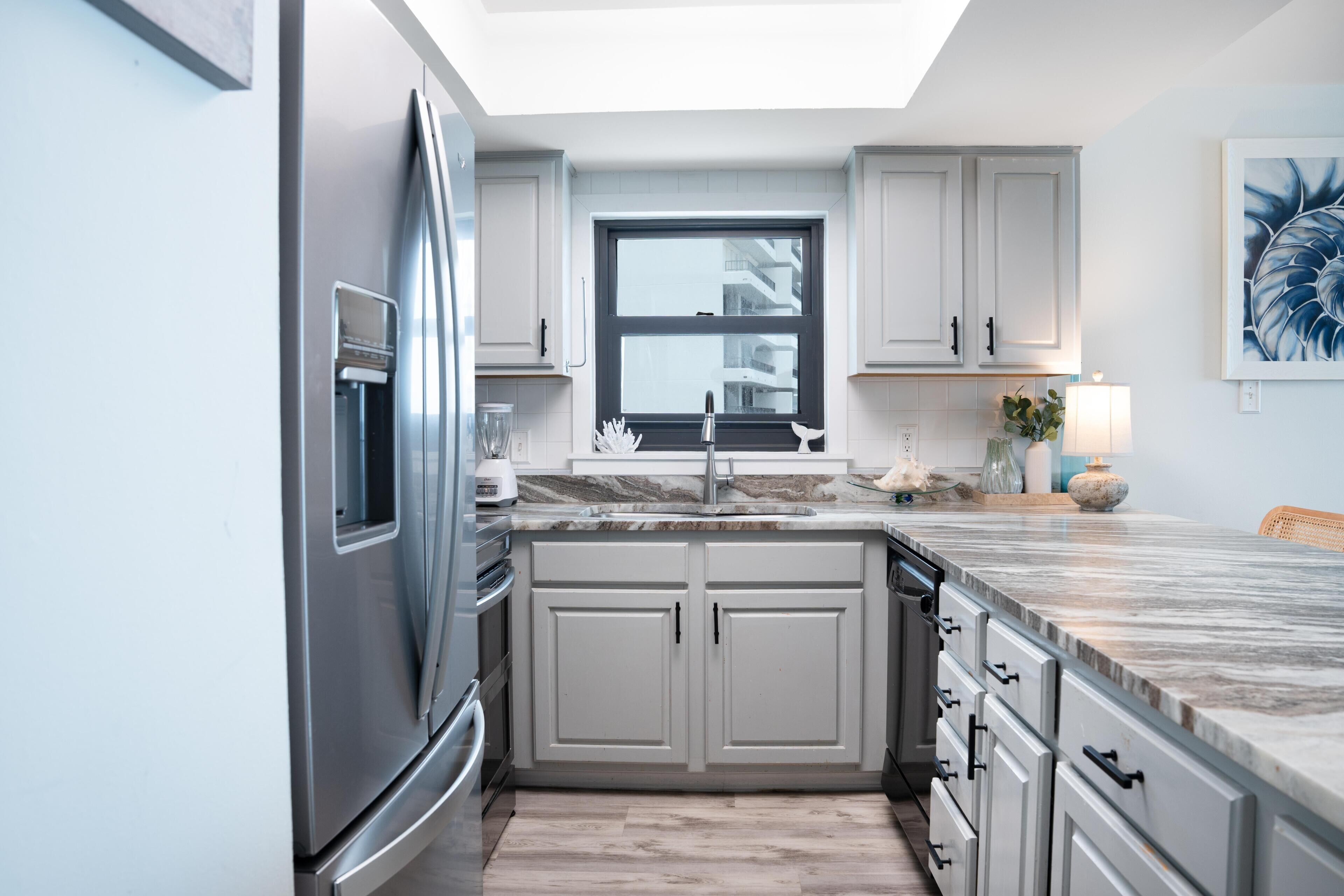 A modern kitchen with white cabinetry, a stainless steel refrigerator, a marble countertop, and a window above the sink.