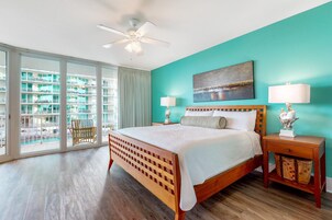 A bedroom with a large bed, two bedside tables, a ceiling fan, and a view of a building through large windows.