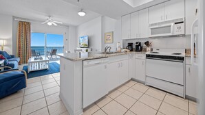 A modern kitchen with white cabinets, a dishwasher, a stove, and a microwave. There is a dining area with a table and chairs, and a view of the ocean through large windows. - Tidewater 503 (Orange Beach)
