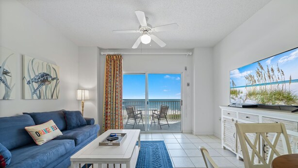 A living room with a blue sofa, white coffee table, and a TV on a cabinet. There's a ceiling fan, a view of the ocean through a sliding door, and a beach scene on the wall. - Tidewater 503 (Orange Beach)