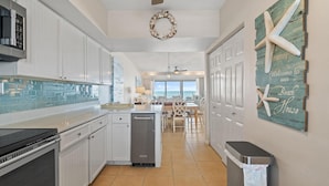 A kitchen with white cabinets, a dishwasher, and a ceiling fan. There is a dining area with a table and chairs, and a view of the outside. - Shipwatch A403 (Pensacola)