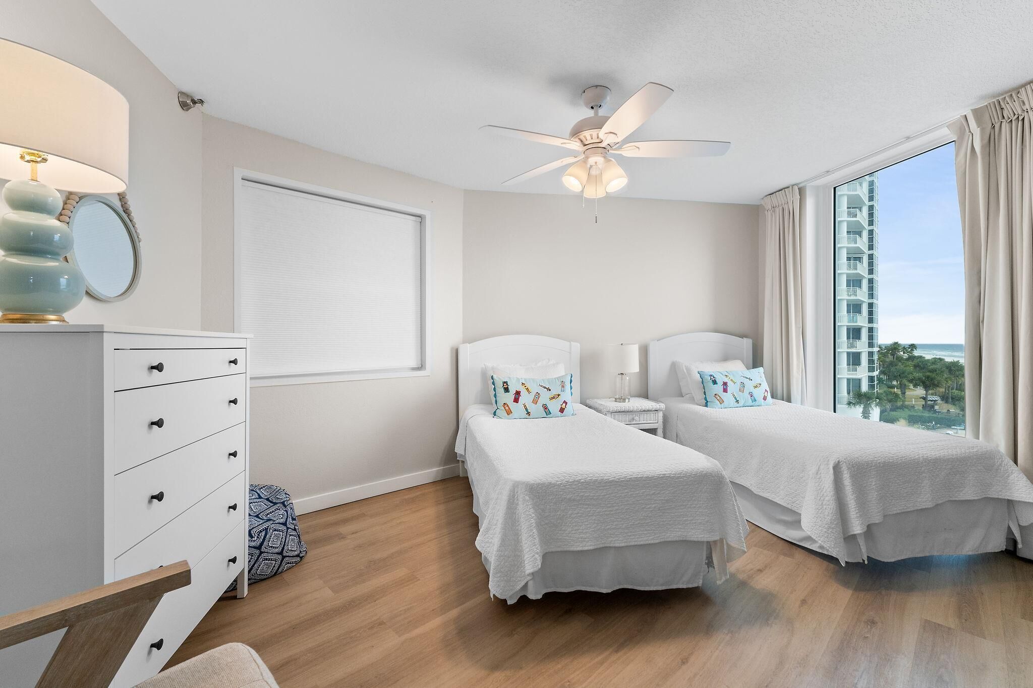 A hotel room with two beds, a ceiling fan, a window with curtains, a dresser, and a bedside table with a lamp.