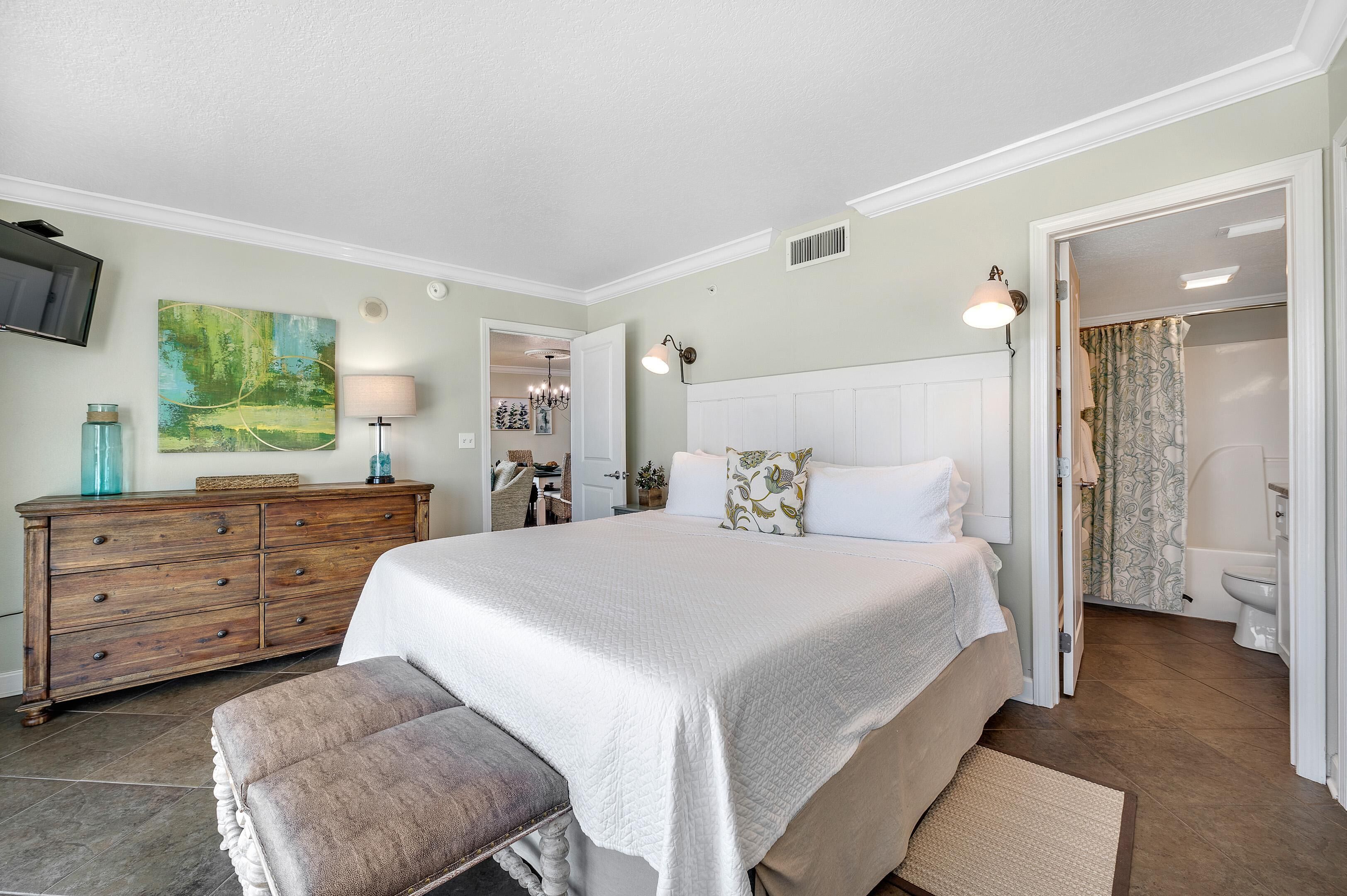 Une chambre à coucher avec un grand lit, une commode en bois, un banc, un tableau et une salle de bain visible à travers une porte ouverte.