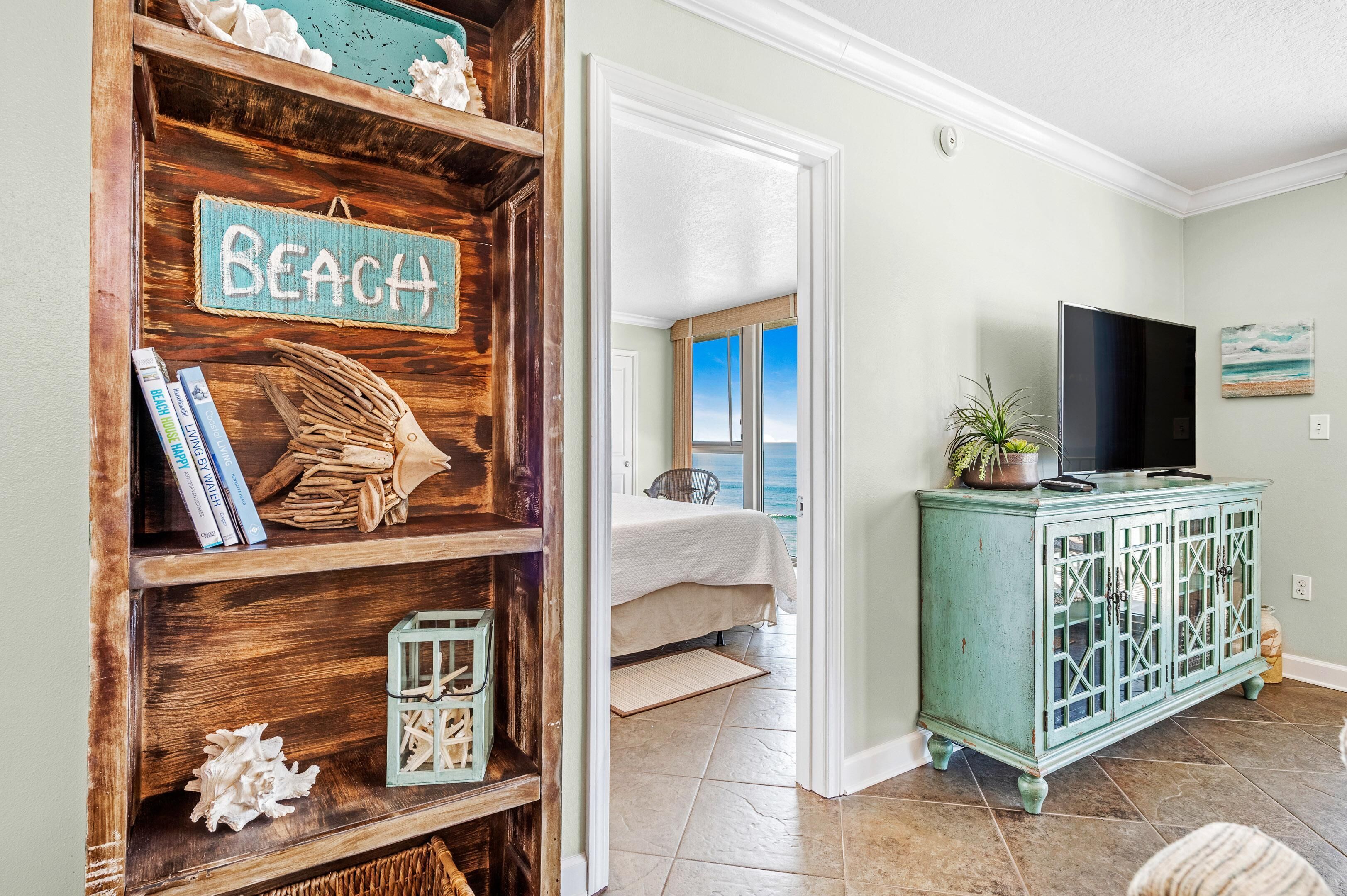 A room with a wooden shelf displaying books and a 'BEACH' sign, a teal cabinet with a TV, and a view of the ocean through a window.