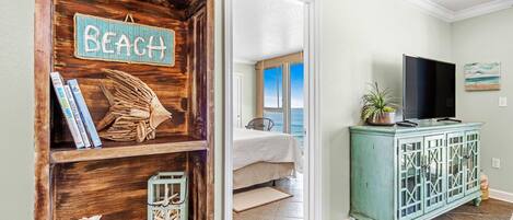 A room with a wooden shelf displaying books and a 'BEACH' sign, a teal cabinet with a TV, and a view of the ocean through a window.