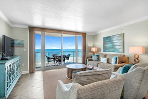 A spacious living room with a large sliding glass door leading to a balcony with a view of the ocean. There is a television on a cabinet, a round coffee table, and a sofa with cushions.