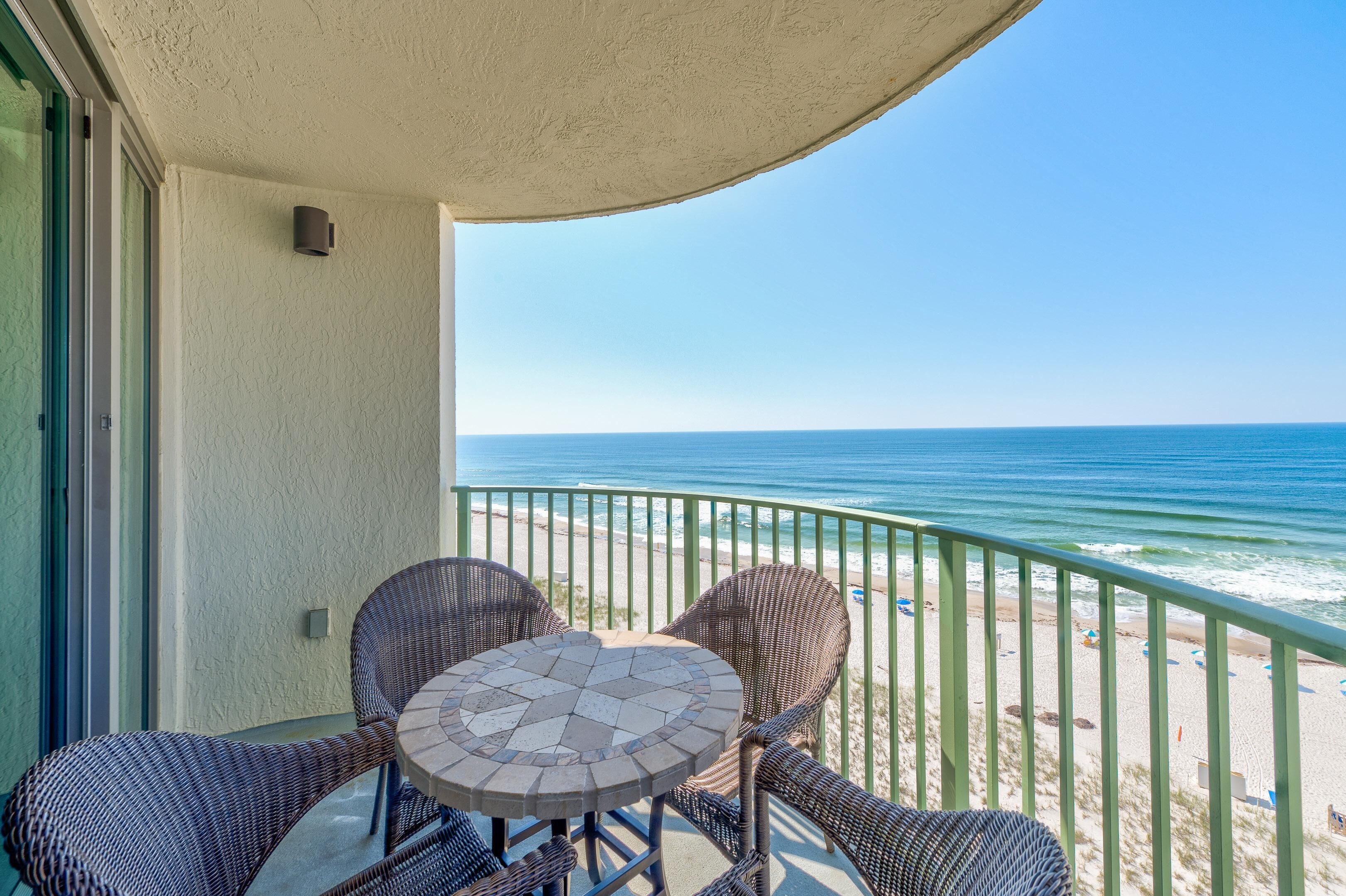 A balcony with wicker chairs and a table, overlooking a beach with the ocean and a clear sky.