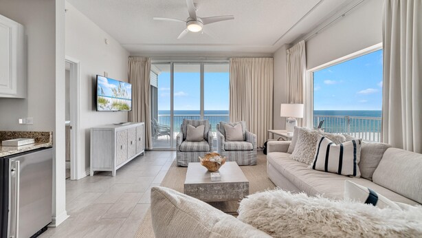 A modern living room with a large window overlooking the ocean, a ceiling fan, and a flat-screen TV mounted on the wall. - Spanish Key 510 (Pensacola)