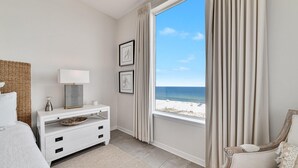 A bedroom with a large window overlooking a beach, a white dresser, a bed, and a chair. - Spanish Key 510 (Pensacola)