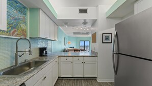 A kitchen with white cabinets, a stainless steel sink, a refrigerator, and a tiled backsplash. - Shipwatch Riverside E1005 (Pensacola)