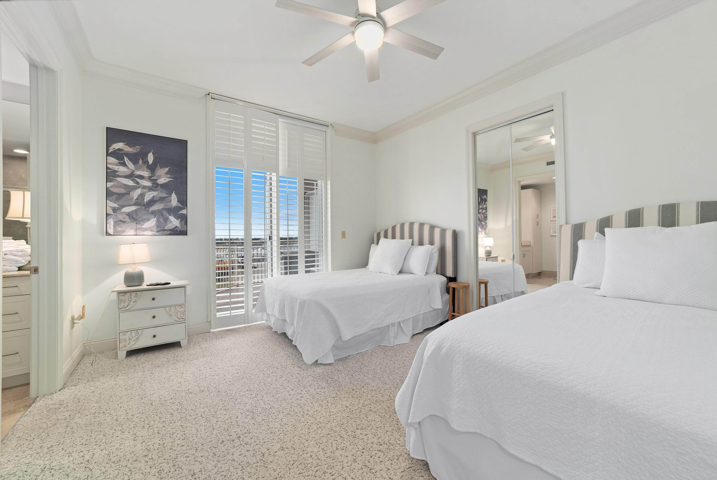 A hotel room with two beds, a ceiling fan, a large window with blinds, a painting on the wall, and a mirror on the door.