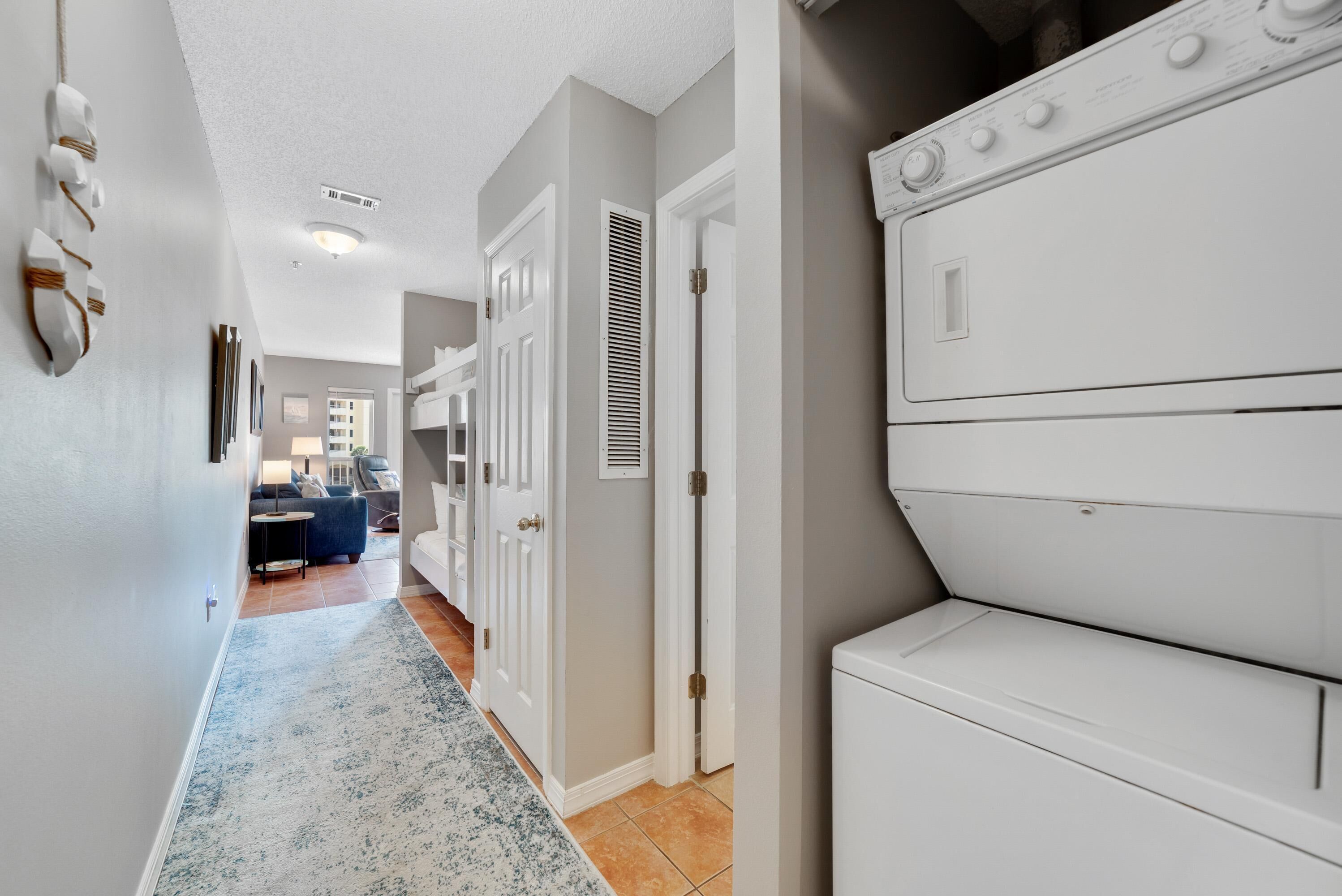 Une buanderie avec un sèche-linge blanc et un lave-linge fixé au mur. Un couloir, orné d’un tapis à motifs bleus, mène à un espace de vie comprenant un canapé et une table basse.